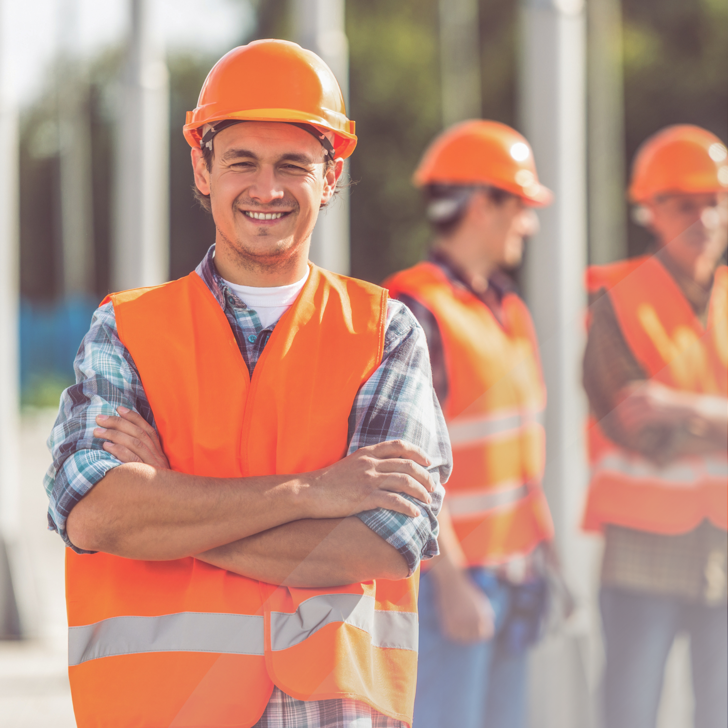 Man stands with arms crossed smiling in hardhat and safety vest, while his co-workers chat behind him.