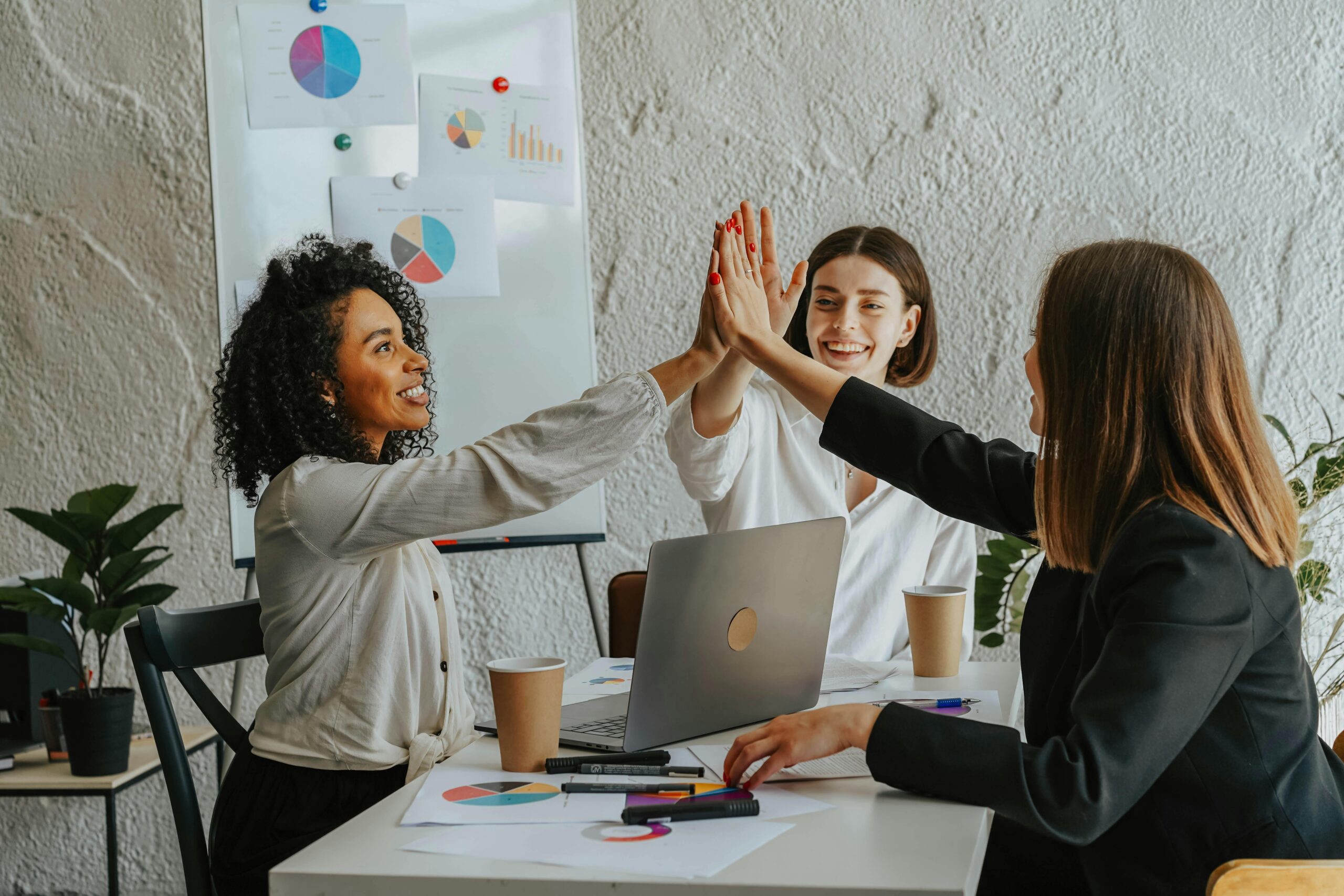 3 people sitting at a desk in a work setting high fiving each other.